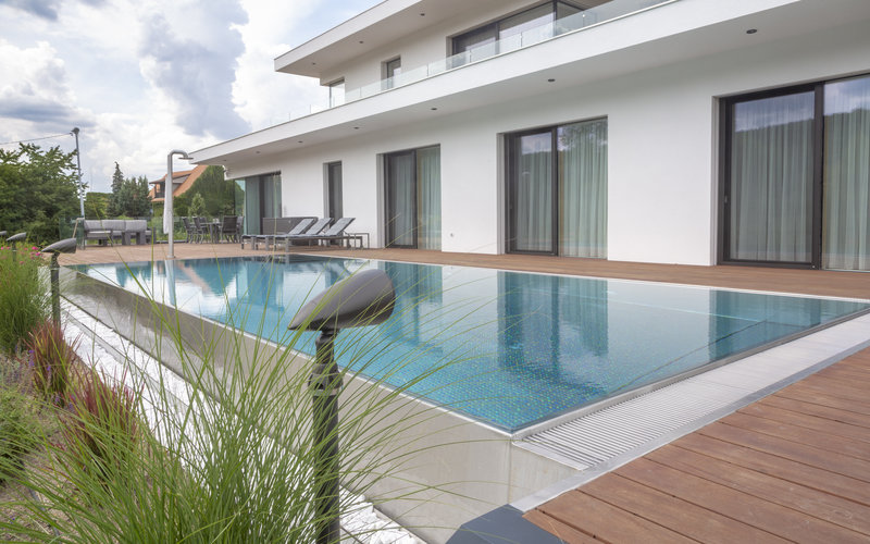 Elegante piscina in acciaio inossidabile con bordo di trabocco a sfioro infinito, acqua turchese e caldo deck in legno accanto a una moderna villa in vetro bianco – circondata da ornamenti erbacei e paesaggio giardino. Piscina in acciaio inossidabile su misura e piscina a sfioro infinito di IMAGINOX per ville private, aree esterne e giardini di lusso. Piscina a sfioro infinito privata, piscina in acciaio inossidabile esterna, piscina con trabocco, piscina con deck in legno, piscina da giardino di lusso, piscina privata – design senza tempo, massima igiene e durabilità per progetti costruttivi esigenti.