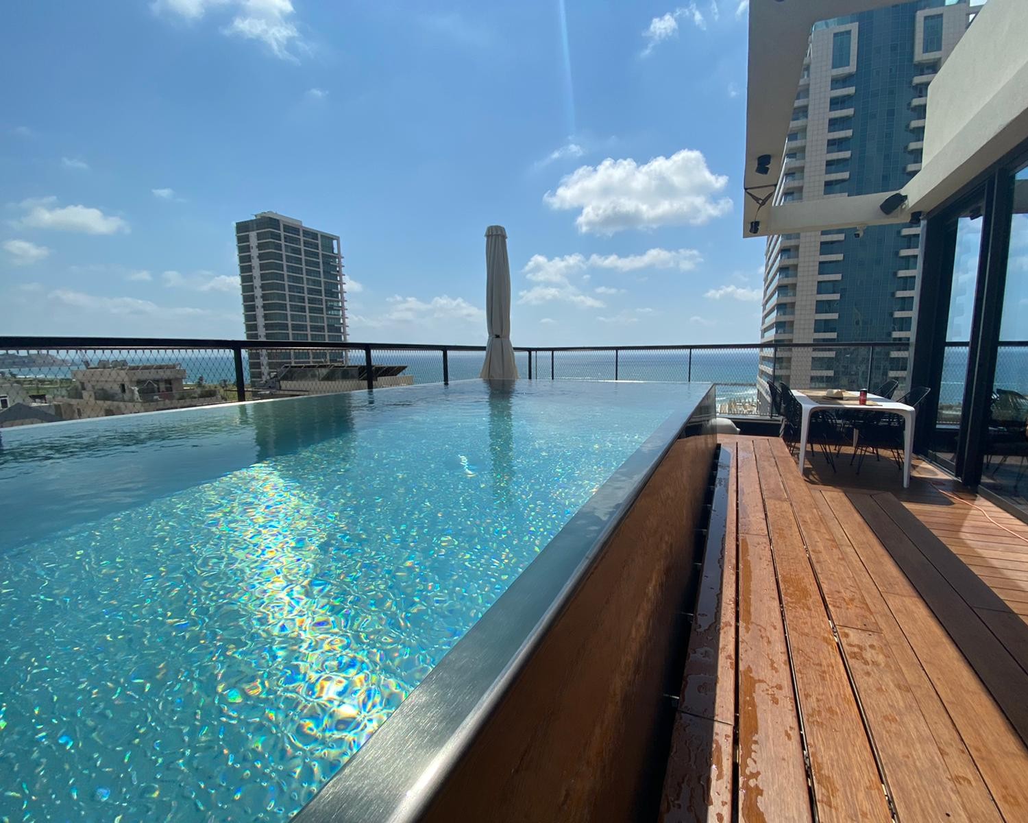 Elegant stainless steel infinity pool with turquoise-blue water and seamless overflow edge on a rooftop terrace with panoramic sea views and skyline – surrounded by warm wooden deck and urban architecture. Custom-made infinity pool by IMAGINOX for rooftop terraces, penthouses, and luxury urban projects. Rooftop terrace infinity pool, rooftop pool, stainless steel overflow pool, infinity pool with sea view, pool on the roof, luxury swimming pool – timeless design, highest precision, and durability for demanding construction projects