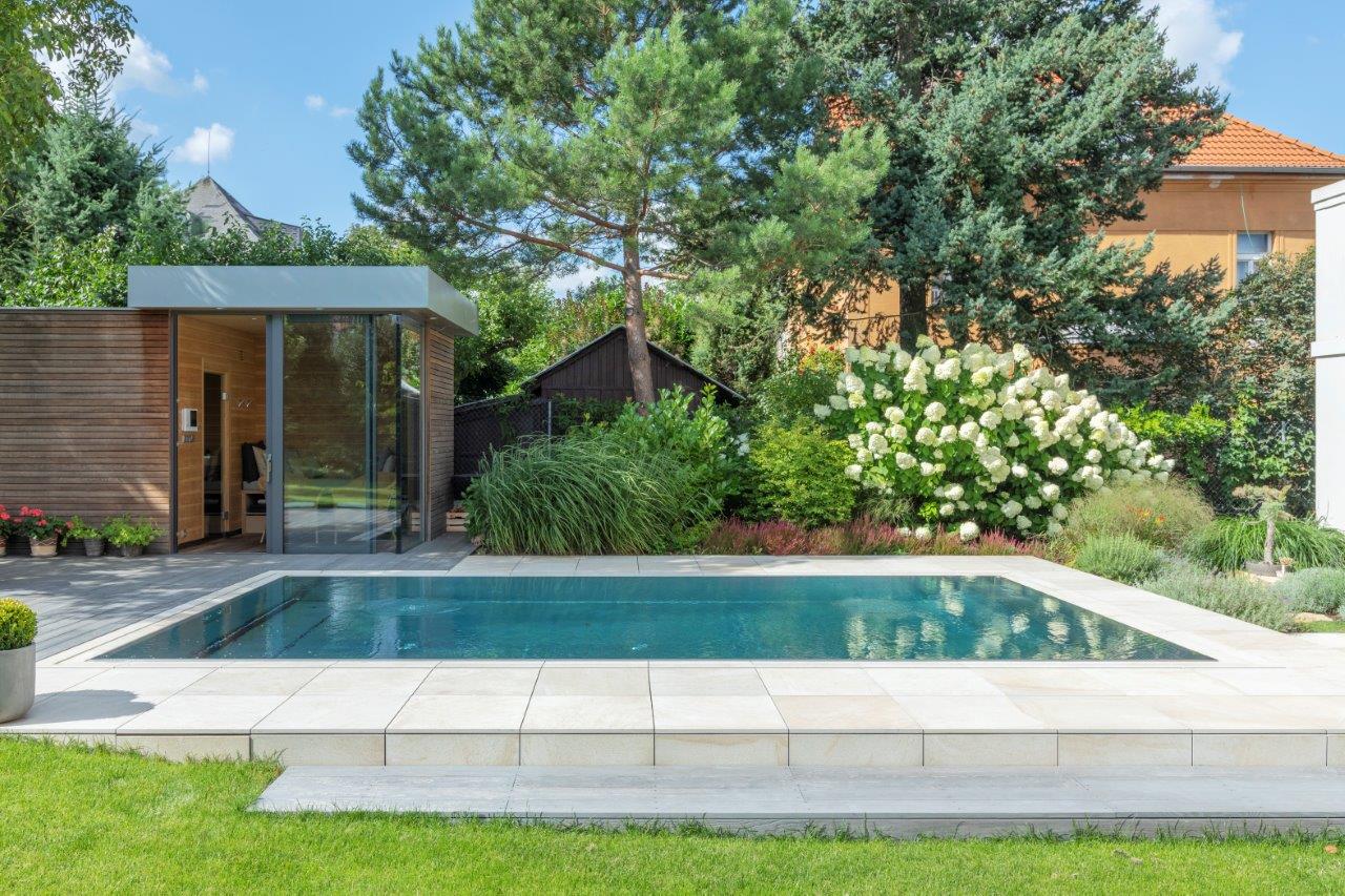 Eleganter Luxuspool aus Edelstahl mit hellem Natursteinrand, türkisblauem Wasser und einem modernen Holz-Saunahaus mit Glasfront – eingebettet in einen üppigen Privatgarten mit weißen Hortensien, Ziergräsern und alten Kiefern. Maßgefertigter Luxuspool von IMAGINOX für private Villen, Stadtgärten und exklusive Außenanlagen. Luxuspool Garten, privater Swimmingpool, Edelstahlpool mit Sauna, Gartenpool Luxus, Pool mit Saunahaus, Außenpool Design – wo Entspannung und Natur zu einem harmonischen Gesamtbild verschmelzen.