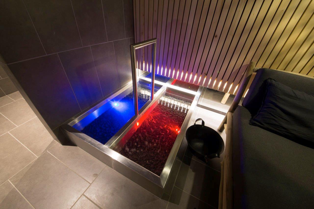 Atmosférický Kneippův chodník z nerezové oceli se dvěma zapuštěnými bazény – modře osvětlený pro studenou vodu, červeně osvětlený pro teplou vodu – obklopený tmavými stěnovými dlaždicemi, teplými dřevěnými lamely a relaxační postelí s polštářem. Vědro na saunový zápar doplňuje klasický saunový rituál. Speciálně vyrobené Kneippovy chodníky od IMAGINOX pro hotely, wellness resorty a soukromé spa oblasti. Kneippův chodník sauna, bazén na brodění teplý studený, Kneippovy chodníky s LED, design Kneippovy instalace, Kneippův chodník nerezová ocel – terapie a atmosféra v jednom.