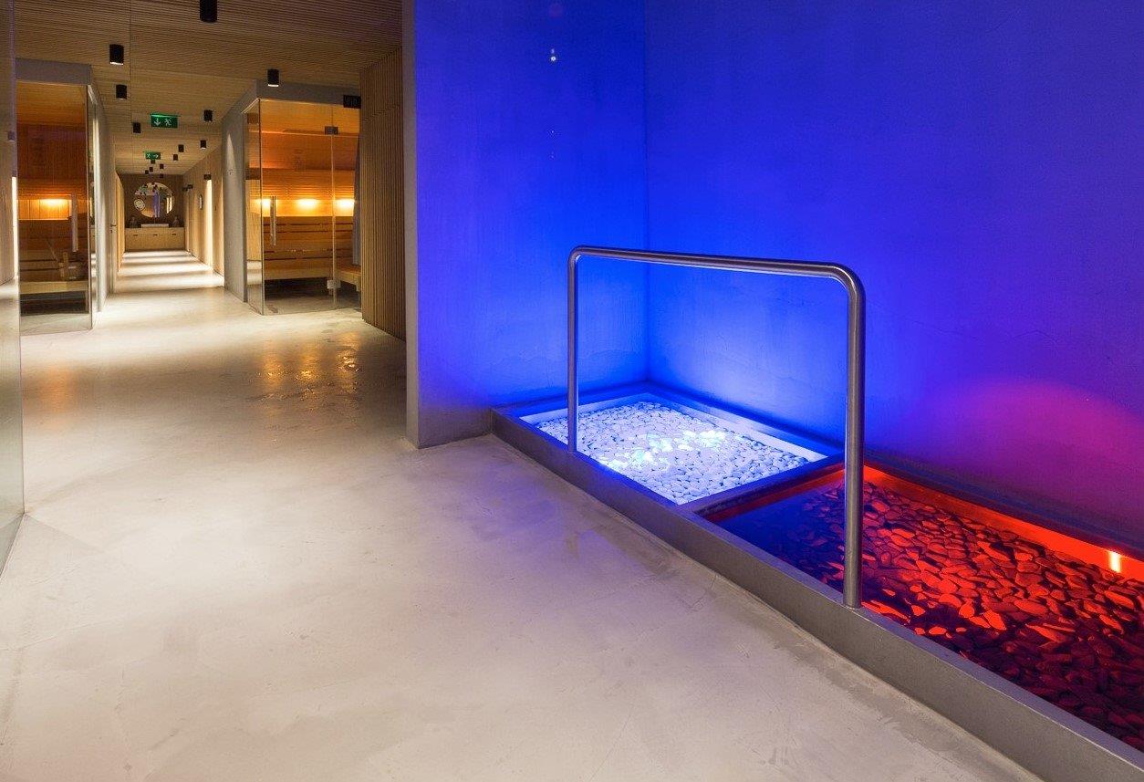 Zapuštěná Kneippova cesta z nerezové oceli se dvěma bazény, bílými a červenými říčními kameny a dramatickým RGB-LED osvětlením v modré a červené barvě – v pozadí dlouhá saunová chodba s teplým dřevěným prostředím. Speciálně vyrobené Kneippovy cesty od IMAGINOX pro hotely, spa centra a komerční wellness oblasti. Kneippova cesta s LED, bazén na chůzi po vodě z nerezové oceli, Kneippovy cesty hotel, design Kneipp-zařízení, Kneippova cesta na míru – zdraví a zážitek v jednom
