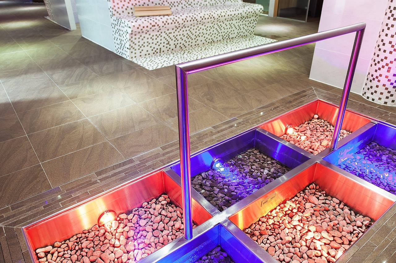 Pohled shora na zapuštěnou Kneippovu cestu z nerezové oceli se čtyřmi bazény, červenými a tmavými říčními kameny a dramatickým RGB-LED osvětlením v červené a modré barvě – Kneippova cesta, která spojuje terapeutický přínos s působivým designem. Speciálně vyrobené Kneippovy cesty od společnosti IMAGINOX pro hotely, wellness resorty a komerční spa oblasti. Pohled shora na Kneippovu cestu, design bazénu na chůzi po vodě, Kneippovy cesty z nerezové oceli, Kneippova cesta s LED, Kneippova zařízení v hotelu – kde zdraví a estetika jdou ruku v ruce.