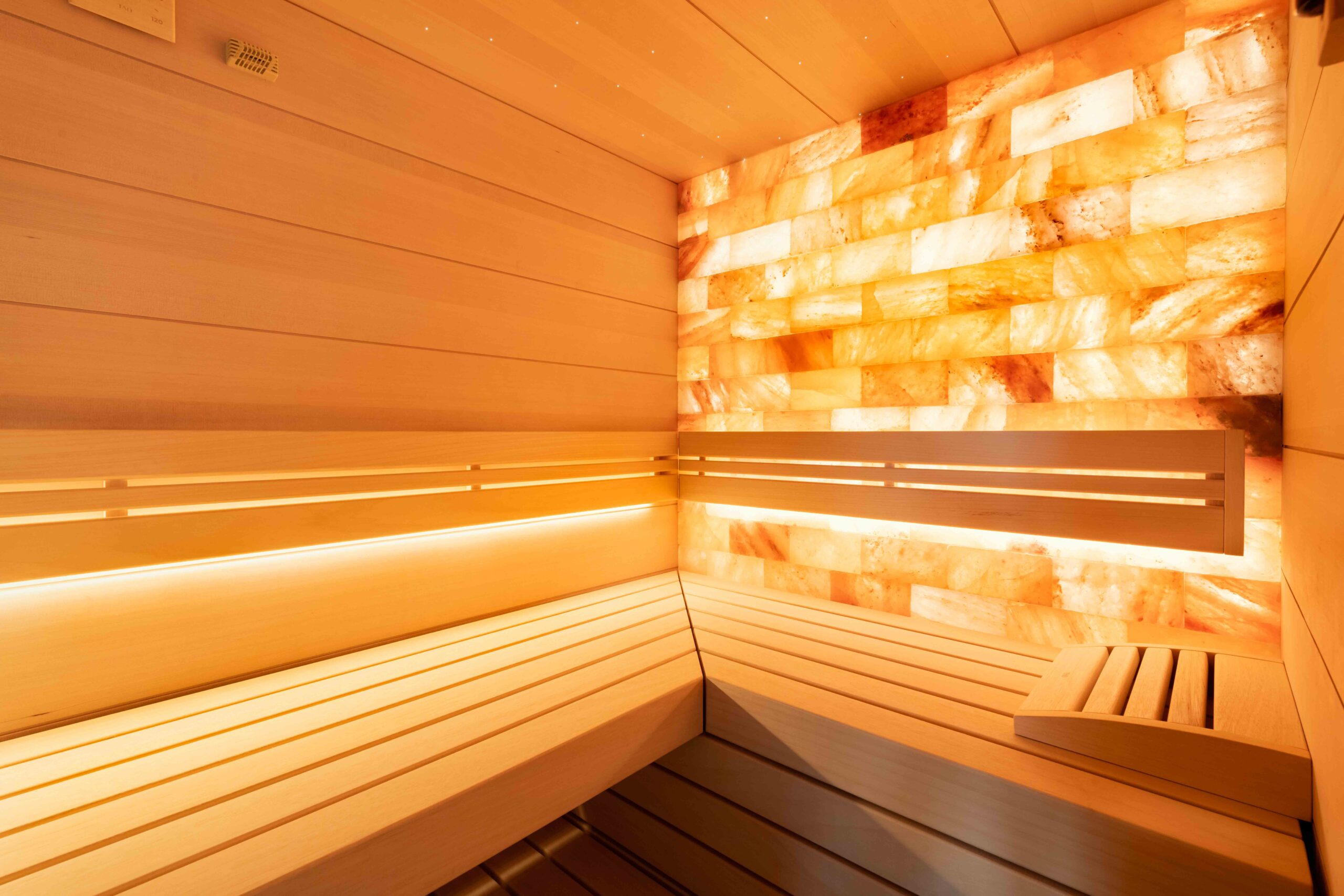 Interior view of an atmospheric custom-made sauna with a glowing Himalayan salt stone wall in warm orange and red tones, light spruce wood benches, warm LED ambient lighting, and a calm, contemplative atmosphere – a sauna that captivates through the natural warmth of the salt stone and the craftsmanship quality of the wood. Custom-made sauna by TAO by IMAGINOX for private villas, hotels, and spa centers. Custom sauna, salt stone sauna, Himalayan sauna, private design sauna, sauna wood LED, sauna for home – relaxation that appeals to all the senses.