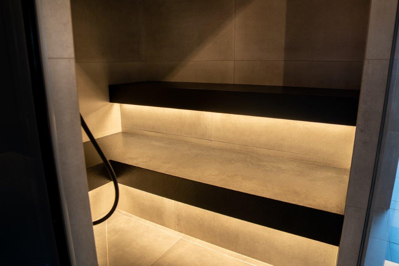 Vnitřní pohled na elegantní parní saunu se třemi víceúrovňovými lavicemi z lehkého přírodního kamene, tmavými dělícími profily a teplým zlatým LED podsvětlením – parní sauna, která se vyznačuje čistými liniemi, vysoce kvalitními materiály a atmosférickým osvětlením. Speciálně vyrobená parní sauna od IMAGINOX pro hotely, spa centra a soukromé wellness místnosti. Vnitřní pohled parní sauna, parní lázeň kamenná lavice, design parní sauna, parní lázeň na míru, moderní parní lázeň – teplo, které vidíte, než ho pocítíte