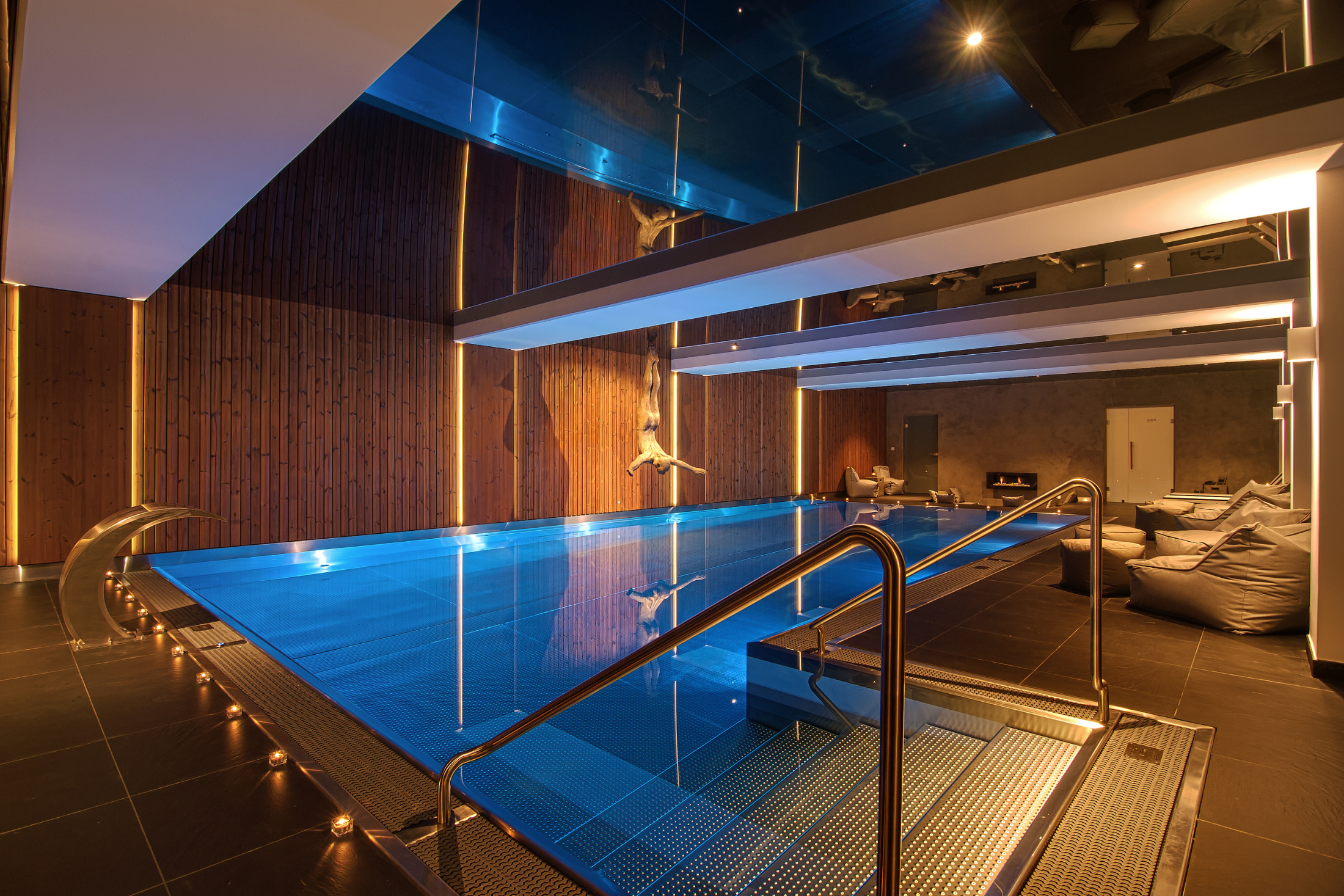 Piscina in acciaio inossidabile su misura di IMAGINOX con illuminazione LED blu in un ambiente elegante, circondata da rivestimento in legno e comodi pouf.