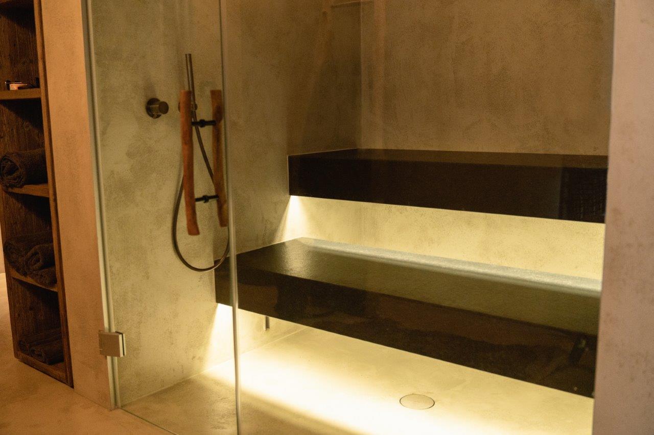 Innenansicht einer eleganten Dampfsauna mit mehrstufigen dunklen Steinbänken, warmem goldenem LED-Unterlicht, Betonoptik-Wänden und einer rahmenlosen Glasfront mit Holzgriff – daneben ein Handtuchregal aus Naturholz und eine Handbrause. Eine Dampfsauna, die durch minimalistische Ästhetik, hochwertige Materialien und stimmungsvolle Beleuchtung eine einzigartige Wohlfühlatmosphäre schafft. Maßgefertigte Dampfsauna von IMAGINOX für Hotels, Spa-Zentren und private Wellnessräume. Dampfsauna Design, Dampfbad Steinbank, Dampfsauna Glasfront, Dampfbad nach Maß, moderne Dampfsauna – Wärme, die man sieht, bevor man sie spürt.