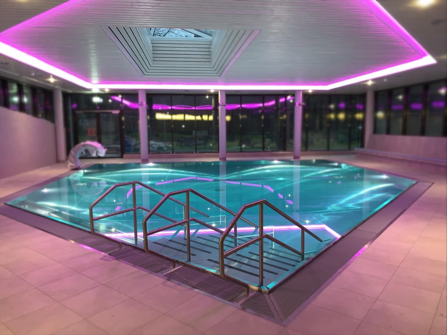 Großzügiger Überlaufpool aus Edelstahl mit türkisblauem Wasser, breitem Edelstahlgeländer und dramatischer pinker RGB-LED-Beleuchtung an Decke und Geländer – eingebettet in einen weitläufigen Hotel-Spa mit rundumlaufender Panorama-Glasfront bei Nacht. Der Überlaufpool wird durch das Farblichtkonzept zum absoluten Erlebnis-Highlight. Überlaufpool Hotel Nacht, Edelstahlpool RGB, Pool LED pink, Hotel Spa Überlaufbecken, Überlaufpool Design – Wellness, die begeistert und in Erinnerung bleibt.