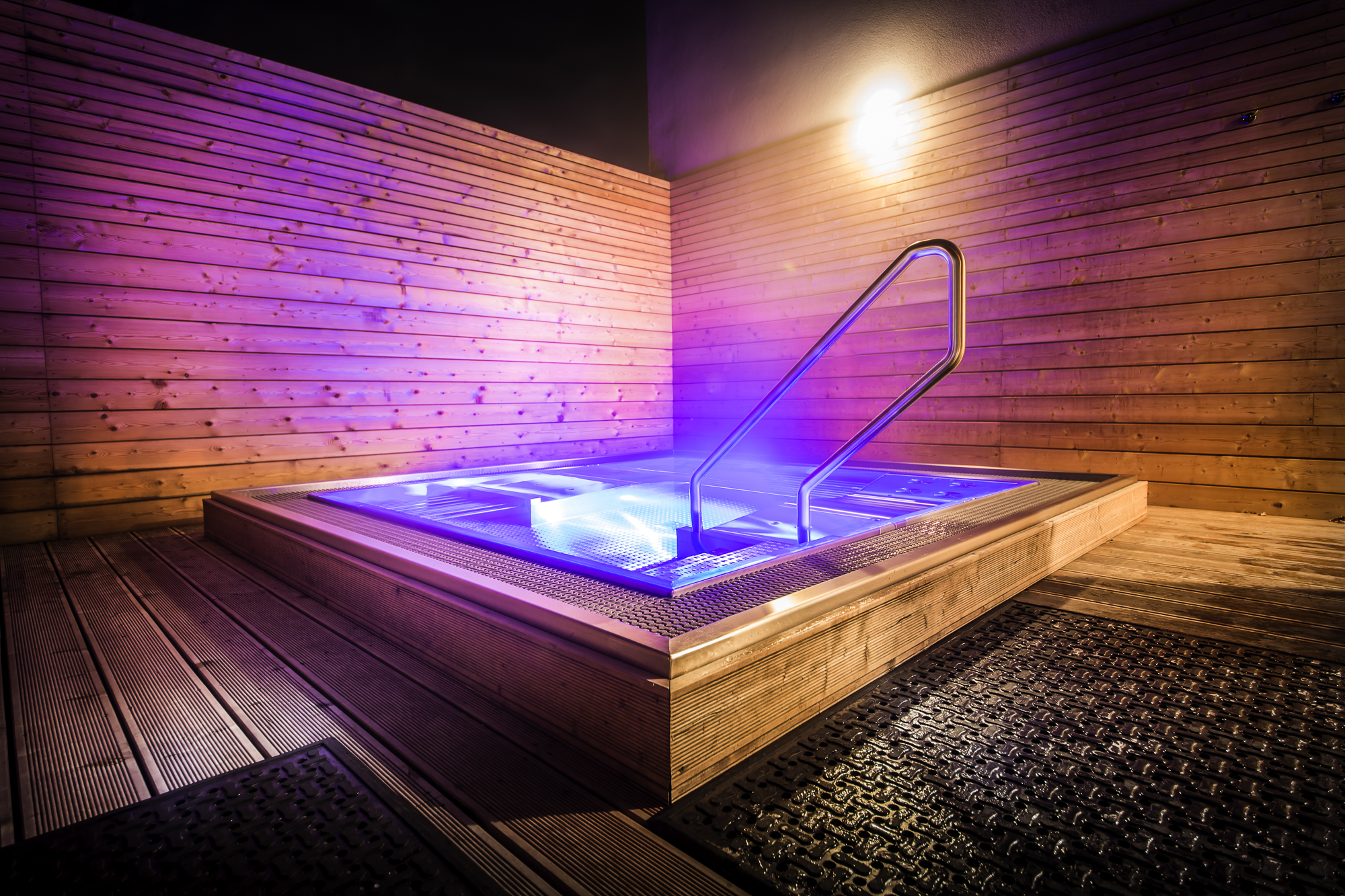 Eingelassener Edelstahl Whirlpool mit leuchtendem Blau-Violett RGB-Licht, Edelstahlgeländer und warmer Holzverkleidung an Wänden und Boden – ein Edelstahl Whirlpool, der durch seine stimmungsvolle Atmosphäre und hochwertige Verarbeitung begeistert. Maßgefertigter Edelstahl Whirlpool von IMAGINOX für Hotels, Wellness-Resorts und gewerbliche Spa-Zentren. Edelstahl Whirlpool Hotel, Whirlpool LED Beleuchtung, Whirlpool Holz Design, Außen-Whirlpool Edelstahl, Wellnessprodukte Hotel – Entspannung und Ästhetik in einem.
