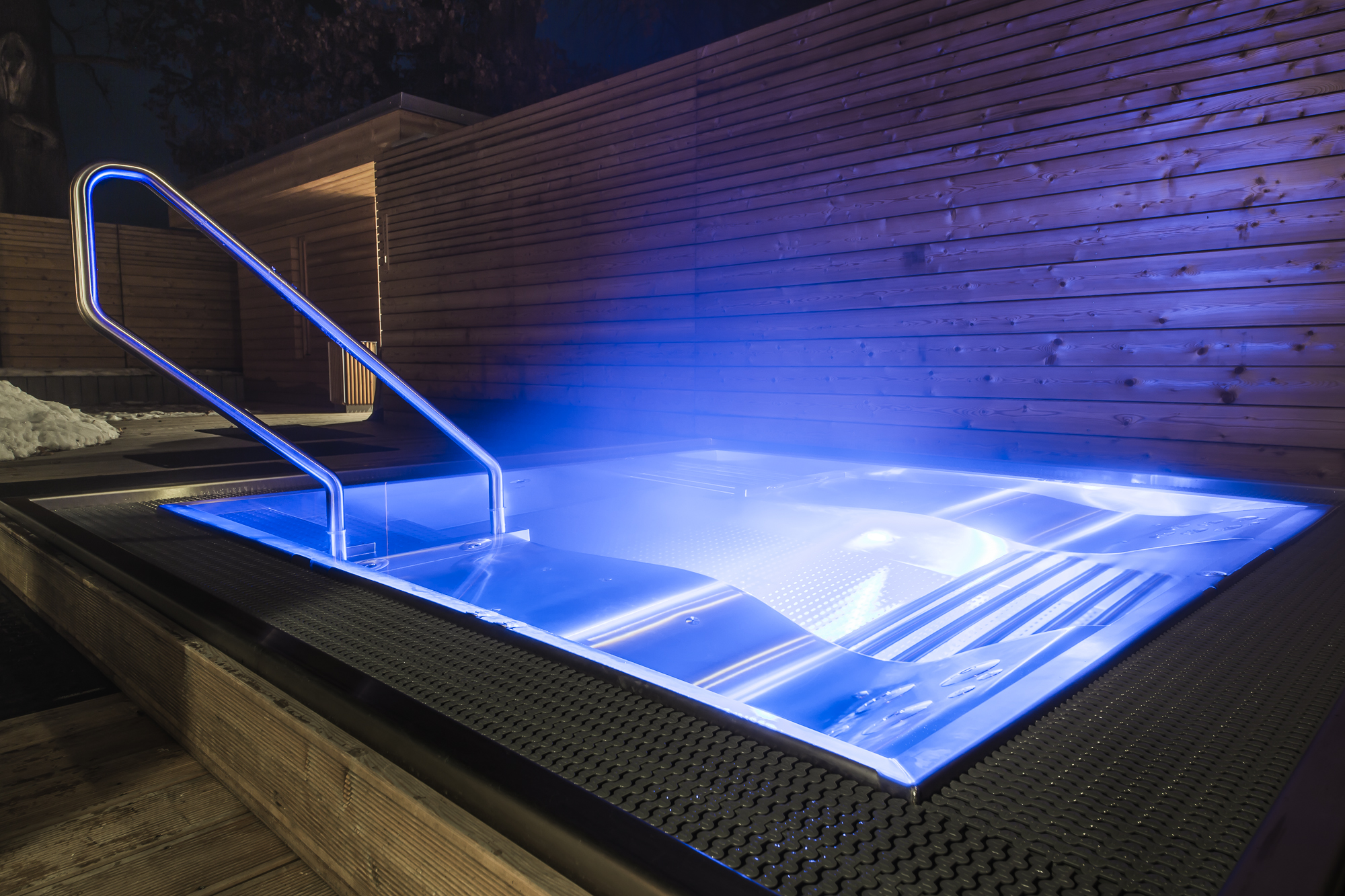 Zářivě modrý vířivý bazén z nerezové oceli v noci se zářícím LED osvětlením, osvětleným zábradlím z nerezové oceli a teplou stěnou dřevěných lamel v pozadí – v zimě obklopený sněhem a noční tiší. Vířivý bazén z nerezové oceli, který svou dramatickou světelnou inscenací a mrazuvzdornou konstrukcí nadchne v každém ročním období a v každém počasí. Vířivý bazén z nerezové oceli na míru od společnosti IMAGINOX pro hotely, wellness resorty a soukromá venkovní zařízení. Vířivý bazén z nerezové oceli v zimě, venkovní vířivý bazén s modrým LED osvětlením, vířivý bazén v noci, vířivý bazén v sněhu, vířivý bazén z nerezové oceli v hotelu – teplo a světlo v nejtmav