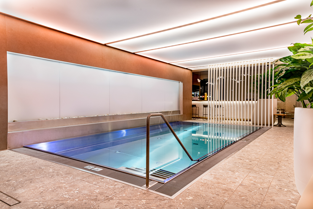 Eingelassener Überlaufpool aus Edelstahl mit türkisblauem LED-Licht, Edelstahlgeländer, weißem Wasserfall-Wandelement und eleganten weißen Lamellen als Raumteiler – eingebettet in einen modernen Hotel-Spa mit warmem Terrakotta-Deckenambiente, Natursteinboden und üppiger Zimmerpflanze. Überlaufpool Hotel, Edelstahlpool Spa Design, Innenpool modern, Pool Wasserfall, Überlaufbecken Hotel – zeitloses Design trifft höchste Qualität