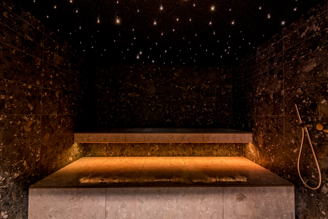 Úchvatná parní sauna s třpytivou hvězdnou oblohou tvořenou světelnými body na tmavém stropě, velkoplošnými tmavými mramorovými stěnami se zlatými vměstky, teplým oranžovým LED podsvětlením pod kamennou lavicí a sprchovou hlavicí v barvě růžového zlata – parní sauna, která svou výjimečnou volbou materiálů a dramatickou světelnou inscenací vytváří jedinečný zážitek. Parní sauna na míru od společnosti IMAGINOX pro luxusní hotely, spa centra a soukromé wellness prostory. Parní sauna hvězdná obloha, parní lázeň tmavý mramor, parní sauna luxus, parní lázeň na míru, hammam design – relaxace pod hvězdným nebem.