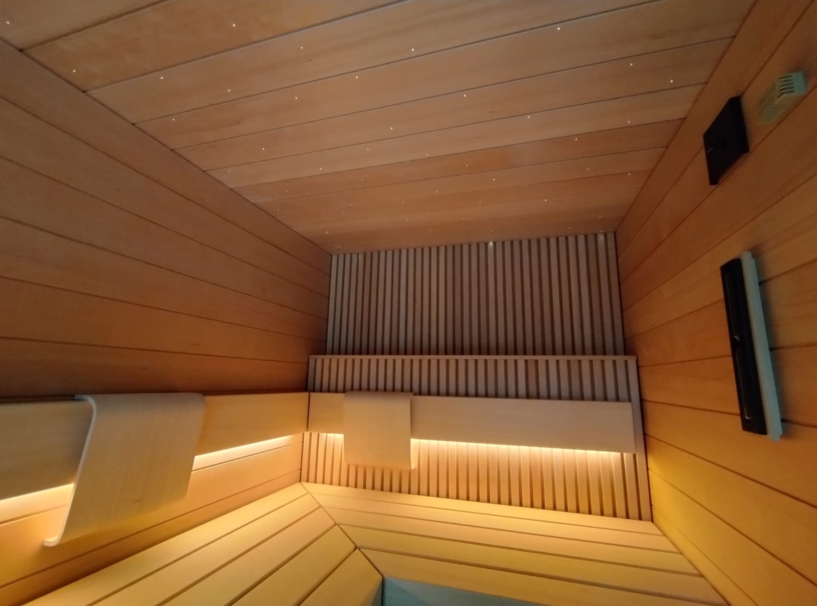 Innenansicht einer außergewöhnlichen Sauna nach Maß mit leuchtendem Sternenhimmel in der Holzdecke, ergonomisch geformten Kopfstützen, warmen LED-Lichtstreifen unter den Bänken und einer eleganten Lamellenrückwand aus hellem Holz – eine Sauna, die durch ihre Liebe zum Detail und die außergewöhnliche Handwerksqualität begeistert. Maßgefertigte Sauna von TAO by IMAGINOX für private Villen, Hotels und Spa-Zentren. Sauna nach Maß, Sauna Sternenhimmel, finnische Sauna Design, Privatsauna Luxus, Sauna Kopfstütze, Sauna LED – Wärme und Komfort auf höchstem Niveau.