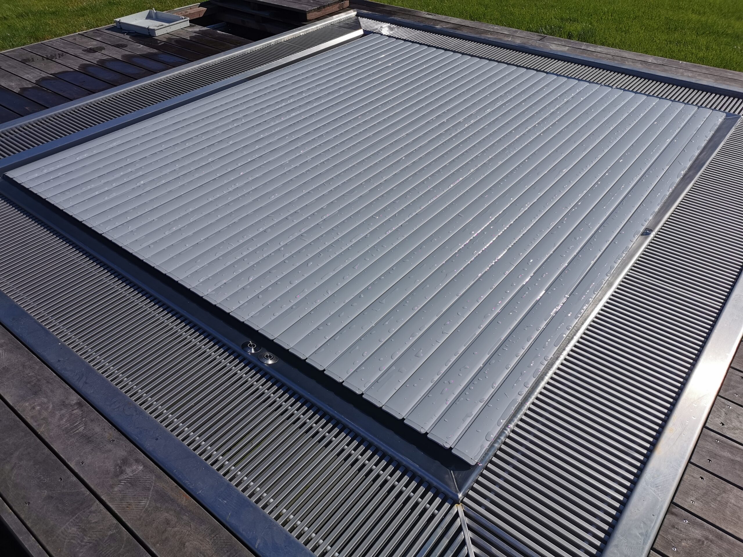 Pohled shora na zcela uzavřený šedý kryt bazénu s přesně vyrobenými PVC lamely – automatické lamelové krytí chrání podkladový nerezový vířivý bazén před nečistotami, listím a ztrátou tepla a dlouhodobě šetří náklady na vytápění a údržbu. Šedý kryt bazénu se harmonicky hodí do dřevěné terasy a při nepoužívání je téměř neviditelný. Zakázkové kryty bazénů a lamelové krytiny od společnosti IMAGINOX pro nerezové vířivé bazény, venkovní bazény a soukromé zahradní areály. Kryt bazénu šedý, lamelové krytí bazénu, automatické krytí bazénu, krytí nerezového vířivého bazénu, lamelové krytí bazénu, ochrana bazénu – praktické, energeticky úsporné a estetické.