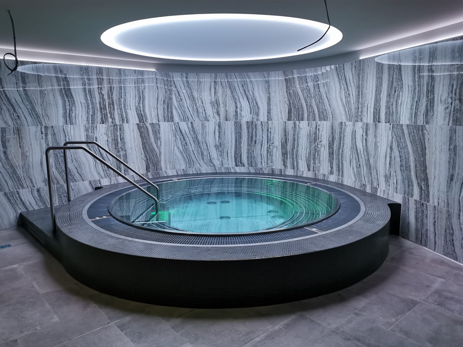 A round, floor-level stainless steel whirlpool with a step and atmospheric green underwater lighting in an elegant wellness room featuring gray-and-white veined marble walls.