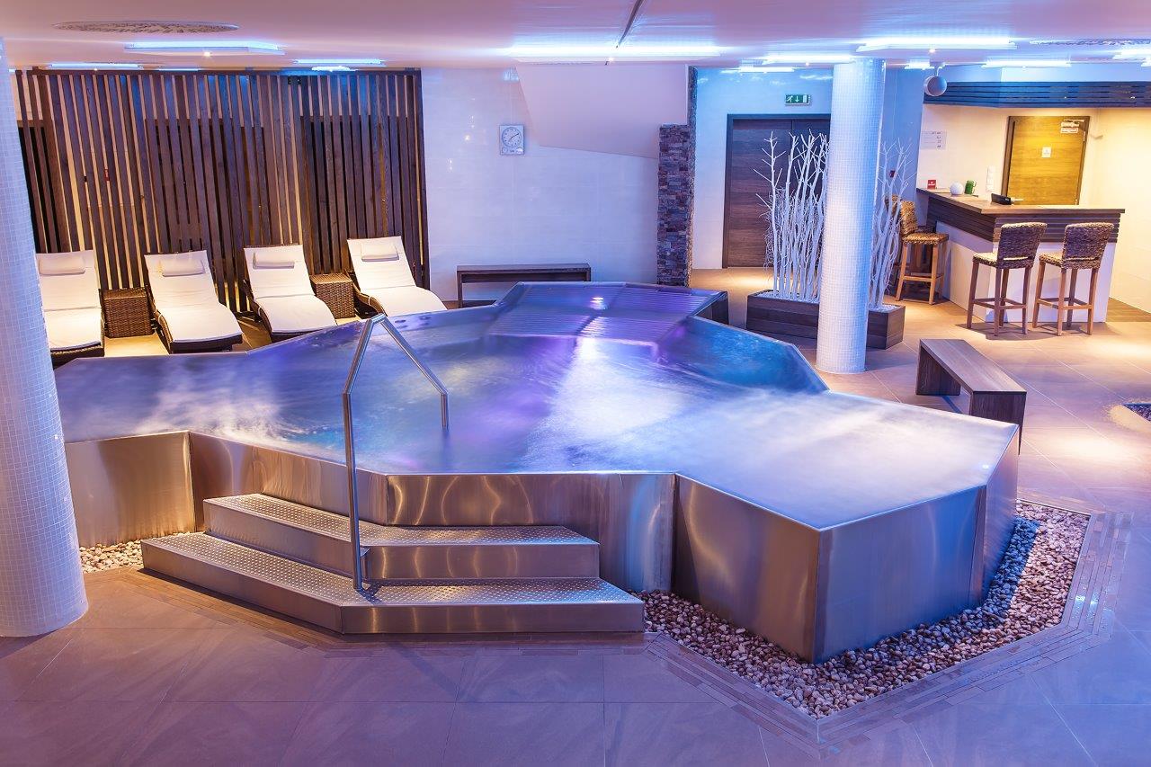 A large, freestanding, custom-made stainless steel hot tub in a striking polygonal shape, featuring illuminated purple water, located in a modern hotel spa area with lounge chairs.