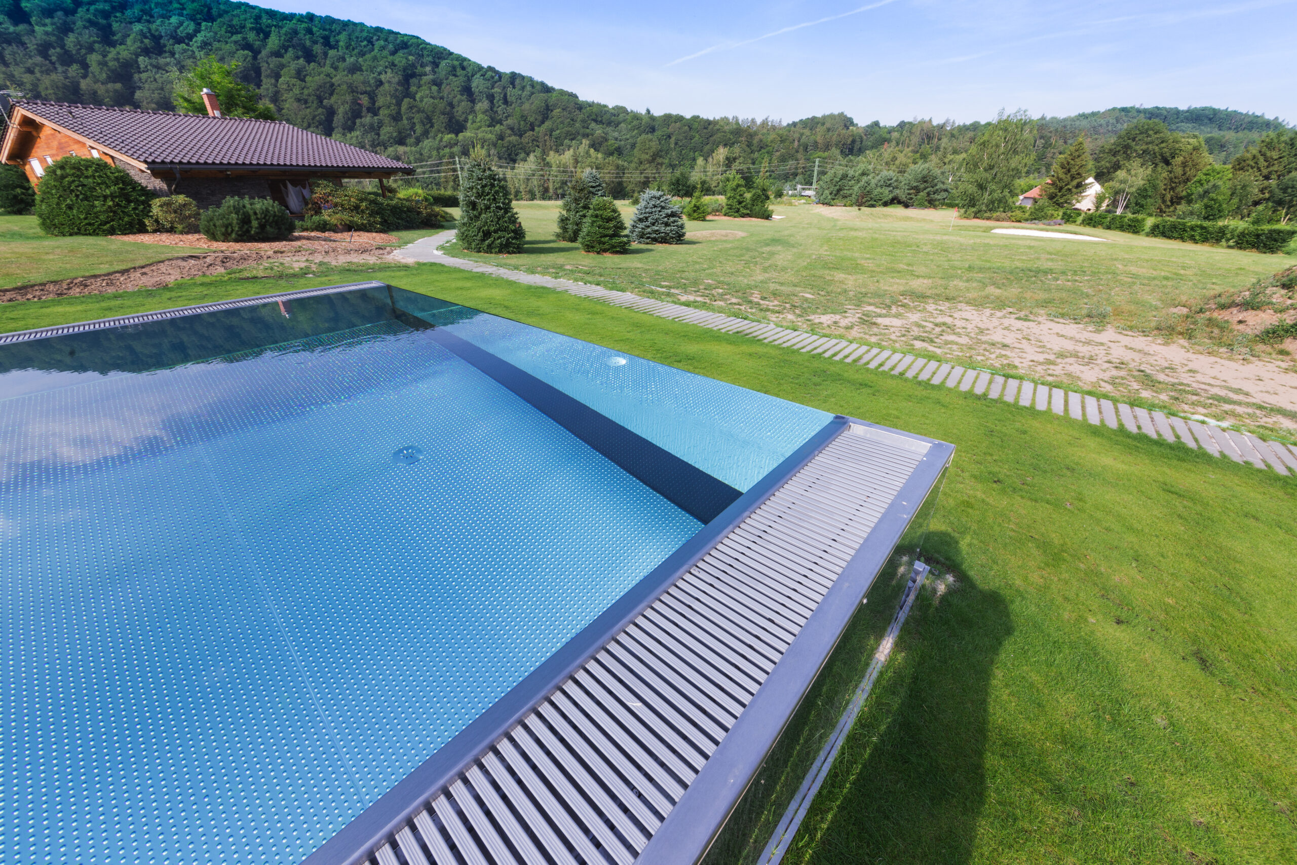 Maßgefertigter Edelstahlpool von IMAGINOX auf einem Hanggrundstück – Luftaufnahme mit Überlaufkante, Gegenstromkanal und Blick auf die Hütte und die grüne, hügelige Landschaft