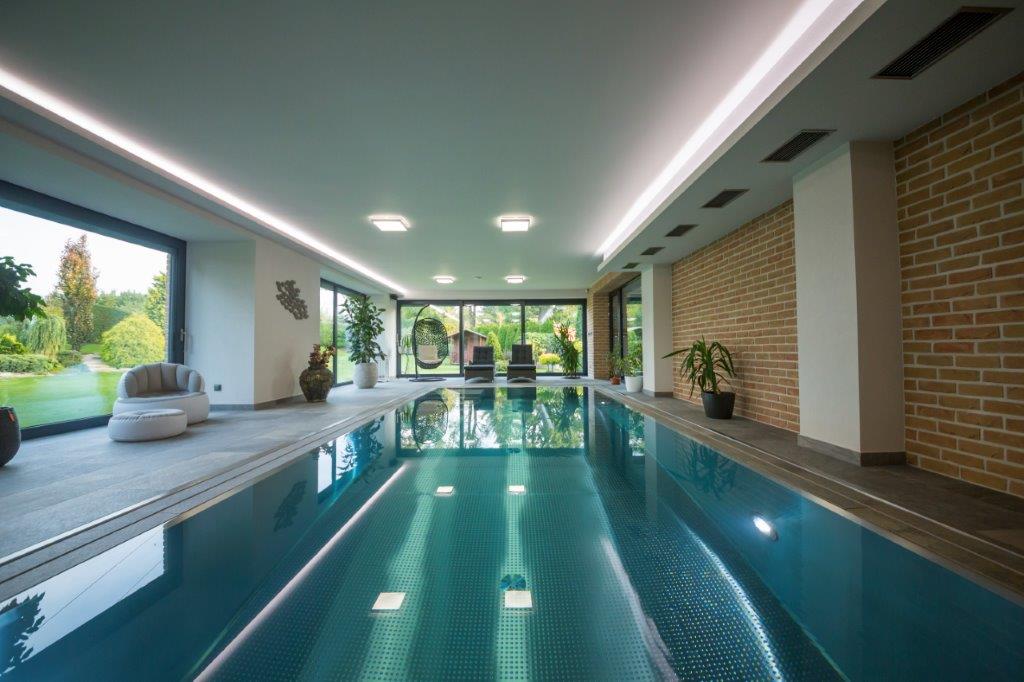 Edelstahlpool von IMAGINOX in einer lichtdurchfluteten Schwimmhalle mit Panoramafenstern, Backsteinwand und Blick in den Garten.