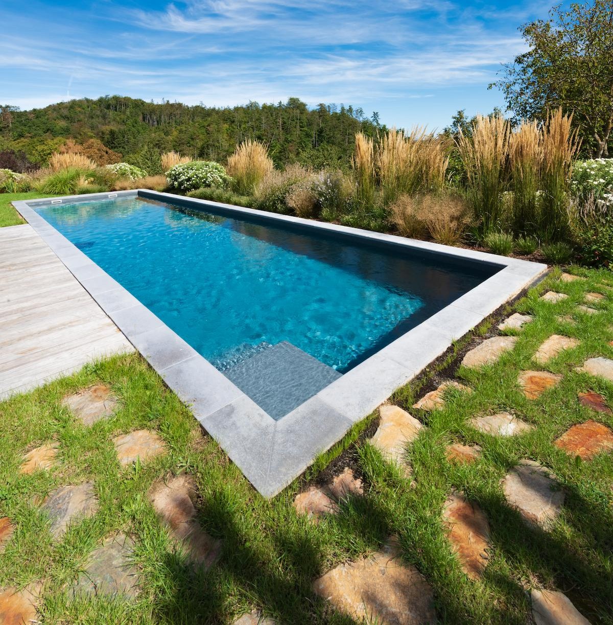 Betonpool von IMAGINOX mit klarem blauem Wasser, eingebettet in einen naturnahen Garten mit Ziergräsern und Trittsteinen.